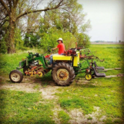 Person wearing a red shirt and white hat smiling at the camera while driving a green and yellow tractor out of a field.